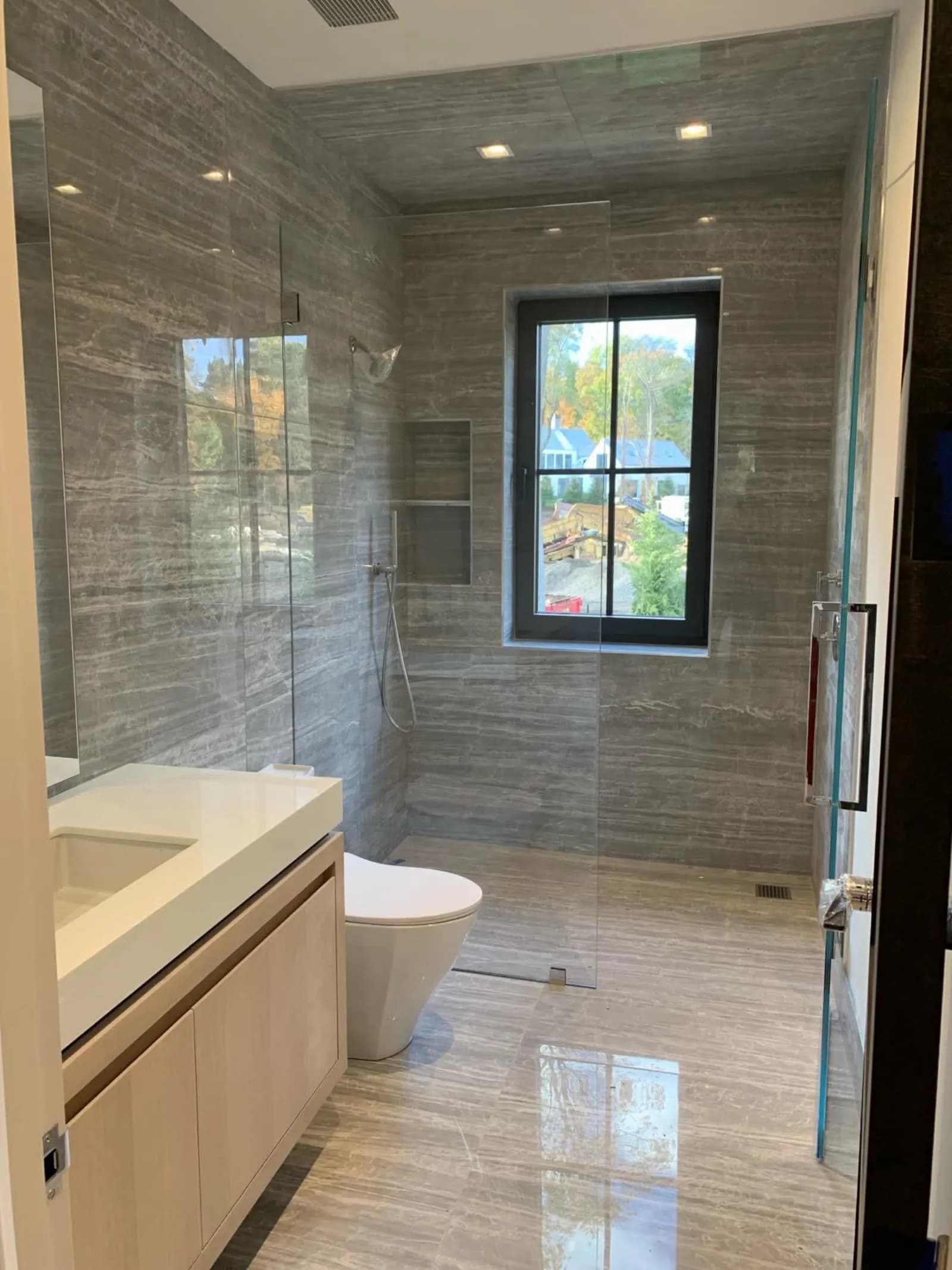 Bathroom with gray stone-look tile walls, frameless glass walk-in shower and light wood vanity